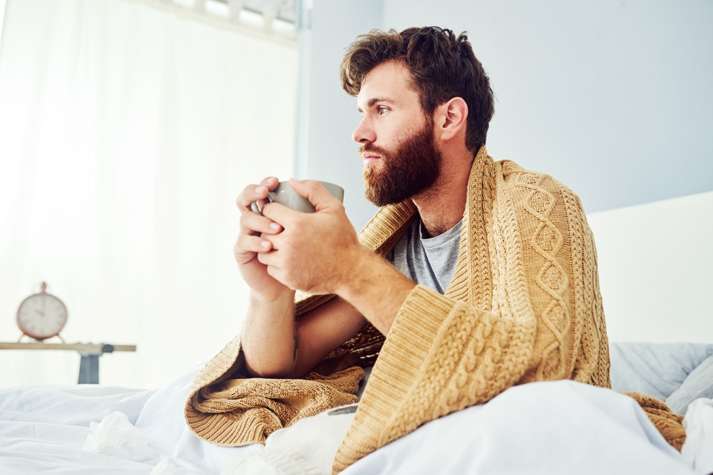 A young man is having a cup of coffee while sitting in bed.