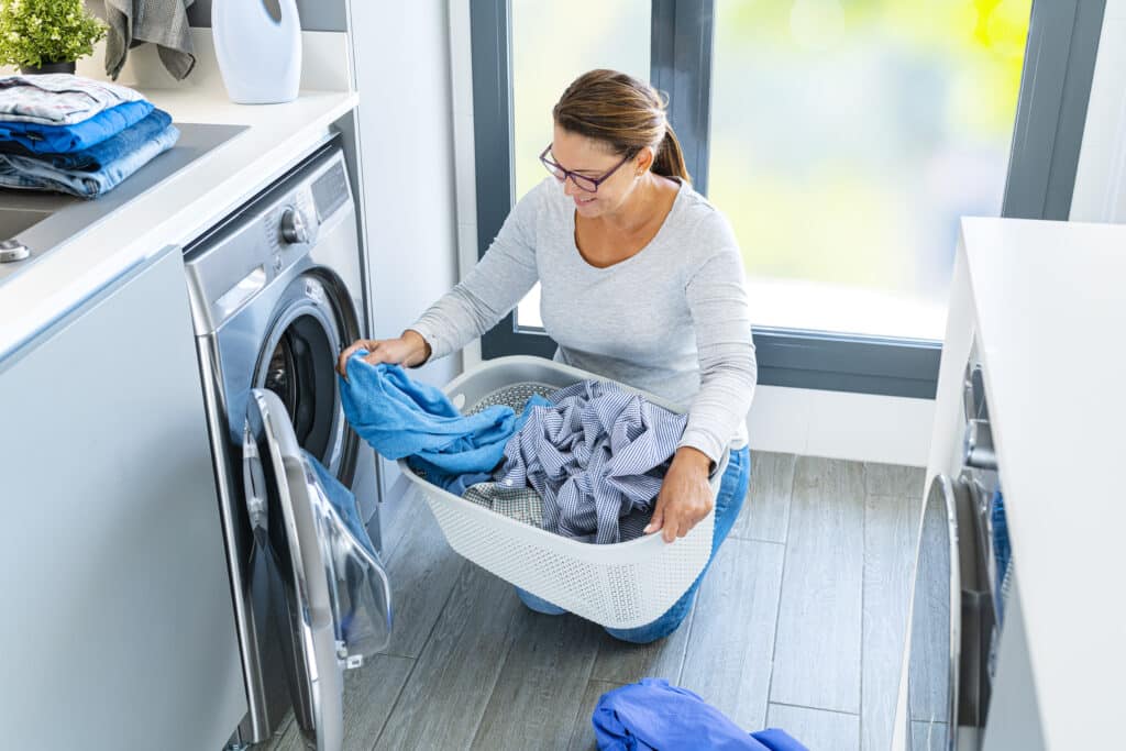 Mature smiling woman loading washing machine with laundry at home.